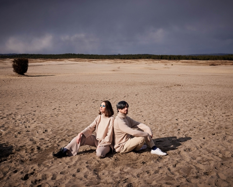 Na zdjęciu kobieta i mężczyzna. Oboje siedzą na piasku plecami do siebie. Mają na sobie beżowa golfy i spodnie. Oboje mają ciemne włosy. Powieki kobiety pomalowane są na niebiesko. Wokół piaski pustyni oraz otaczający ją las.